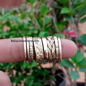 Peut inclure: Un ensemble de sept bagues en métal doré présentant diverses textures, notamment des anneaux lisses, un motif tressé et des gravures florales. Le texte BalajiJewellersGifts est visible sur les bagues. Elles sont disposées côte à côte.