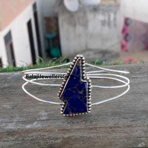 May include: A silver-toned cuff bracelet with a large, irregular-shaped, deep blue stone at the center. The stone has gold-colored veining. The bracelet has three parallel bands and a beaded border around the stone. The text "BalajiJewellersGi" is visible.