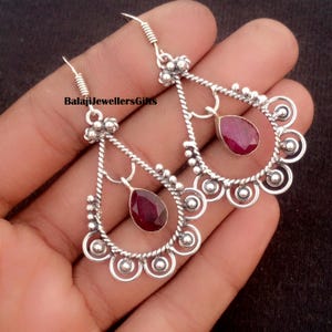 May include: Silver-toned dangle earrings with teardrop-shaped, deep red gemstones. The earrings feature a decorative design with a twisted rope pattern and small circular accents. The text "BalajiJewellersGifts" is visible.