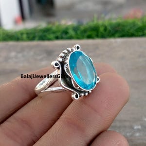 Pode incluir: Um anel de prata com uma grande pedra preciosa oval azul claro. O anel tem uma cravação detalhada com elementos decorativos ao redor da pedra. O texto "BalajiJewellersGifts" é visível.