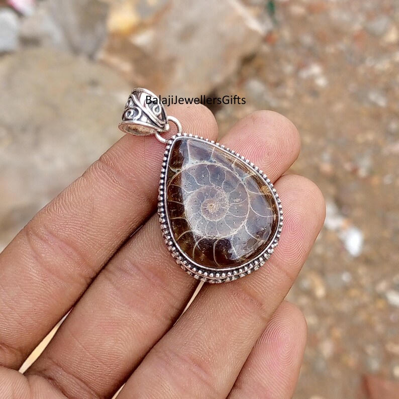 May include: A teardrop-shaped pendant featuring a polished, brown ammonite fossil with a spiral pattern. The fossil is set in a silver-colored metal frame with a decorative bail. The text "BalajiJewellersGifts" is visible.
