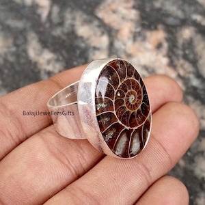 Puede incluir: Un anillo de plata con un amonita fosilizada ovalada. La amonita muestra un patrón espiral detallado en tonos marrones y beige. La banda del anillo es de plata pulida. El texto "BalajiJewellersGifts" es visible en el anillo.