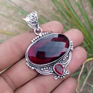 May include: A silver pendant with a large, faceted, oval-shaped, deep red gemstone as the focal point. A smaller, teardrop-shaped red gemstone is below. The pendant has ornate silver detailing and the text "BalajiJewellersGifts".