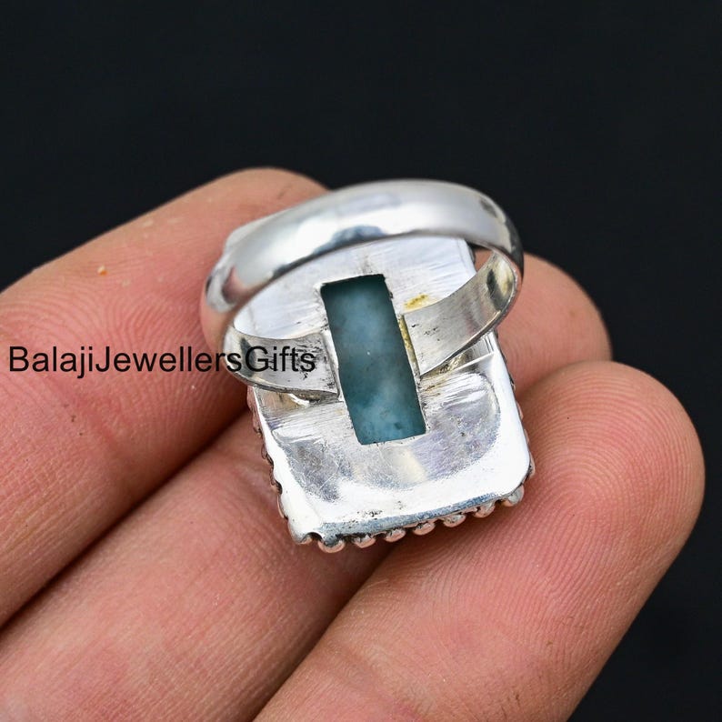 K&ouml;nnte beinhalten: Ein silberner Ring mit einem rechteckigen blauen Edelstein, der in einer silbernen Fassung sitzt. Der Ring hat einen perlenbesetzten Rand und ein glattes Band.
