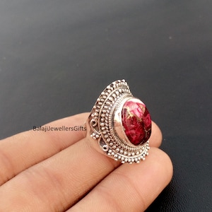 以下が含まれることがあります： 楕円形の赤と金の石が付いたシルバーリング。リングは石の周りとバンドに詳細でテクスチャ加工されたデザインが施されています。「BalajiJewellersGifts」というテキストがリングの側面に表示されています。