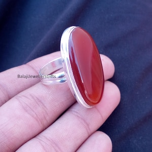 Op de afbeelding: Een zilveren ring met een grote, ovale, gepolijste rode edelsteen. De ring heeft een eenvoudige band en een bezel setting. De tekst "BalajiJewellersGifts" is zichtbaar op de ring.
