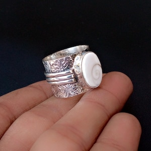 Puede incluir: Un anillo de plata con una piedra ovalada blanca. El anillo tiene una banda giratoria y está decorado con patrones florales. El anillo se sostiene en una mano sobre un fondo oscuro. El texto "RajiJewellersGifts" es visible en el anillo.