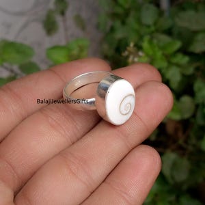 Pode incluir: Um anel de prata com uma pedra branca ovalada grande com um desenho em espiral. O anel é segurado em uma mão contra um fundo verde desfocado. O texto "BalajiJewellersGifts" é visível no anel.