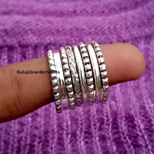 Puede incluir: Un conjunto de anillos de plata con varios diseños, incluyendo bandas texturizadas y lisas. Los anillos se muestran en un dedo sobre un fondo morado. El texto "BalajiJewellersGifts" es visible.