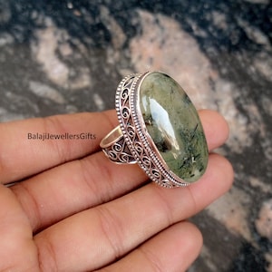 Könnte beinhalten: Ein ovaler Silberring mit einem grünen Edelstein, der dunkle Einschlüsse aufweist. Der Ring hat einen dekorativen Filigranrand. Der Text "BalajiJewellersGifts" ist sichtbar. Der Ring wird in einer Hand gehalten.