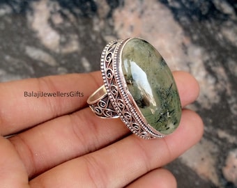Puede incluir: Anillo ovalado de plata con una piedra verde con inclusiones oscuras. El anillo tiene un borde de filigrana decorativo. El texto "BalajiJewellersGifts" es visible. El anillo se sostiene en una mano.
