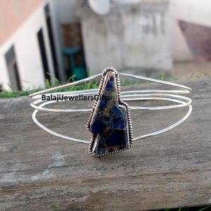 May include: A silver-toned bracelet with a dark blue and gold stone centerpiece. The stone is irregularly shaped and set in a textured silver frame. The bracelet has three parallel bands. The text "BalajiJewellersGifts" is visible.