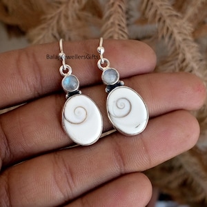 May include: A pair of silver earrings featuring a white oval-shaped pendant with a spiral design. Each earring has a small, round, blue gemstone above the pendant. The earrings are held in a person's hand.