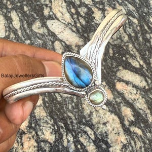 May include: A silver bracelet with a braided design and two iridescent labradorite stones. The bracelet is open and has a curved shape.