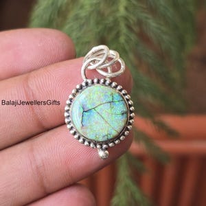 Puede incluir: Un colgante redondo con una fianza plateada y un bisel engastado con un ópalo colorido. El ópalo tiene una mezcla de colores verdes, azules y blancos con un patrón agrietado. El colgante está sujeto por una mano.