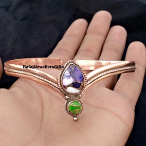 Op de afbeelding: Koperkleurige armband met een druppelvormige paarse en witte steen en een kleine groene steen. De armband heeft de tekst "BalajiJewellersGifts" erop.