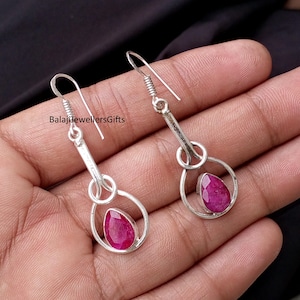 May include: A pair of silver earrings with a teardrop-shaped, deep red gemstone set within a circular frame. The earrings have a hook closure and a decorative spiral detail. The text "BalajiJewellersGifts" is visible.