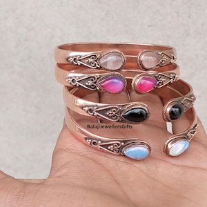 May include: Copper-colored metal cuff bracelets with teardrop-shaped gemstones in pink, black, and blue. The bracelets have an ornate design near the gemstone settings. The text "BalajiJewellersGifts" is visible.