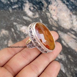 May include: A large, oval-shaped ring with a faceted, amber-colored gemstone. The ring has an ornate, silver-colored filigree setting. The text "BalajiJewellersGifts" is visible on the ring.