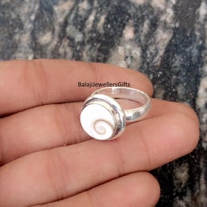 May include: A silver ring featuring a large, round, white stone with a spiral pattern. The ring has a simple band and a bezel setting. The text "BalajiJewellersGifts" is visible.