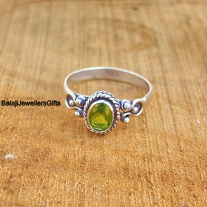 Pode incluir: Um anel de prata com uma gema oval verde engastada em uma banda decorativa. O anel tem um design de corda em torno da pedra e pequenos detalhes decorativos nas laterais.