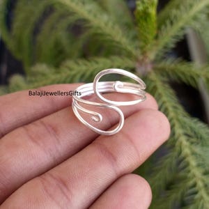 May include: A silver-colored ring with a unique, open design. The ring features a swirling, abstract shape with multiple bands and decorative curves. The ring is shown against a blurred green background. The text "BalajiJewellersGifts" is visible.