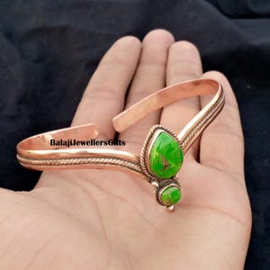 Puede incluir: Un brazalete de cobre con un diseño de alambre trenzado a lo largo de la banda y dos piedras verdes en forma de lágrima con vetas de cobre. El texto BalajiJewellersGifts está impreso en la superficie metálica del brazalete abierto.