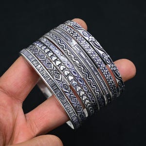 May include: A collection of silver bangles featuring detailed patterns, such as floral, heart, and geometric motifs. The bangles are stacked, displaying diverse textures and designs. The jewellery is presented against a dark backdrop.