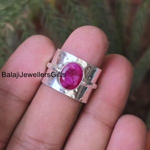 Peut inclure: Une bague en argent avec une grosse pierre précieuse rose vif de forme ovale. La bague a un large anneau texturé et le texte "BalajiJewellersGifts". La pierre précieuse est au centre, sertie dans un sertissage à lunette.