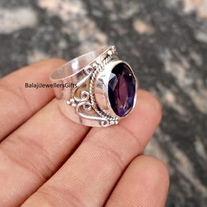 May include: A silver ring featuring a large, oval, deep purple gemstone. The ring is intricately designed with decorative elements around the stone and on the band. The text "BalajiJewellersGifts" is visible.