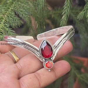 May include: A silver bracelet with a teardrop-shaped, deep red gemstone and a smaller, round orange gemstone. The bracelet has a decorative rope-like design along the edges and is held in a hand.