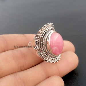 Puede incluir: Un anillo de plata con una gema rosa ovalada grande. El anillo tiene un diseño ornamentado y texturizado con pequeños detalles de cuentas alrededor de la gema y a lo largo de la banda. El texto "BalajiJewellersGifts" es visible.