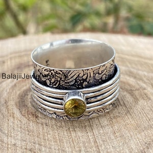 May include: A silver spinner ring with an ornate floral design on the top band and a yellow gemstone. The ring has a textured, layered design with a spinning center band. The ring is on a wooden surface.