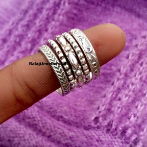 Puede incluir: Un conjunto de anillos de plata con diversos diseños decorativos, incluyendo diseños florales y de cuentas. Los anillos están apilados en un dedo sobre un fondo morado. El texto "BalajiJewellers gift" es visible.
