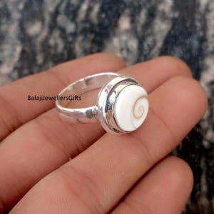 May include: A silver ring with a white, circular stone displaying a spiral pattern. The ring is held in a hand and has a polished finish. The text "BalajiJewellersGifts" is visible on the ring.