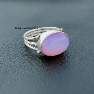 May include: A silver ring featuring a large, oval, pale pink gemstone. The ring has a multi-band design with small decorative spheres. The text "BalajiJewellersGifts" is visible.
