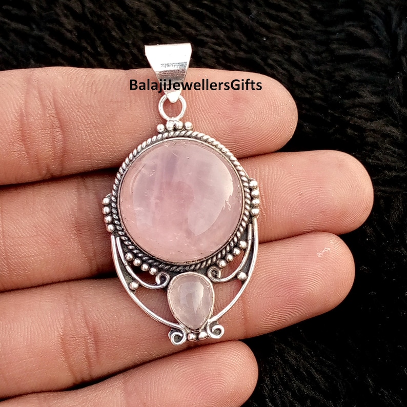 May include: A silver pendant featuring a large, round, pale pink rose quartz gemstone and a smaller, teardrop-shaped stone beneath. The pendant has an intricate silver setting. The text "BalajiJewellersGifts" is visible at the top of the pendant.