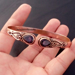 May include: Copper cuff bracelet with two teardrop-shaped, dark purple gemstones. The bracelet features ornate, detailed metalwork around the gemstones and the text "BalajiJewellersGifts".