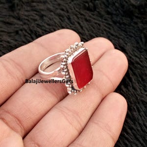 May include: A silver ring with a rectangular, deep red gemstone. The ring has a textured border around the stone and a simple band. The text "BalajiJewellersGifts" is visible on the ring.