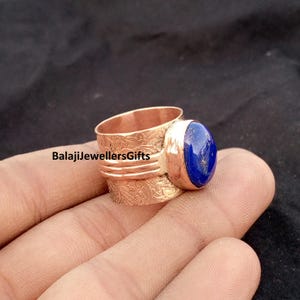 Peut inclure: Une bague en cuivre dotée d'un large anneau avec un motif floral en relief et une pierre précieuse ovale en lapis-lazuli bleu lisse sertie dans une monture. Le texte BalajiJewellersGifts est imprimé sur la surface de l'anneau en métal.