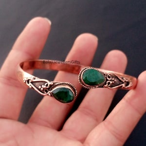 May include: Copper-colored cuff bracelet with two pear-shaped, emerald-green gemstones. The bracelet has an open design with decorative filigree accents. The text "BalajiJewellersGifts" is visible on the bracelet.