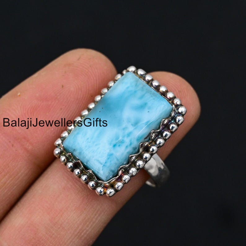 K&ouml;nnte beinhalten: Ein silberner Ring mit einem blauen und wei&szlig;en Edelstein, der in einer Perlenfassung sitzt. Der Ring wird an einem Finger getragen.