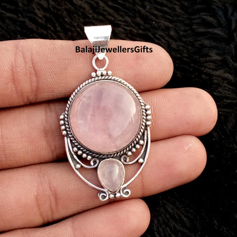 May include: A silver pendant featuring a large, round, pale pink rose quartz gemstone and a smaller, teardrop-shaped stone beneath. The pendant has an intricate silver setting. The text "BalajiJewellersGifts" is visible at the top of the pendant.