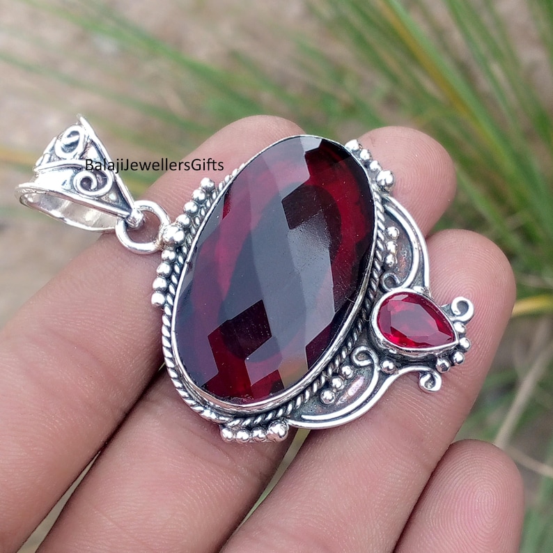 May include: A silver pendant featuring a large, faceted, oval-cut, deep red gemstone, accented by a smaller, teardrop-shaped red stone. The pendant has ornate silver detailing and a bail for a chain. The text "BalajiJewellersGifts" is visible.