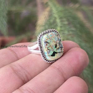 Puede incluir: Anillo de plata con una piedra de ópalo rectangular. El ópalo presenta una mezcla de colores verdes, blancos y negros con una superficie texturizada. El anillo tiene un borde trenzado en forma de cuerda.