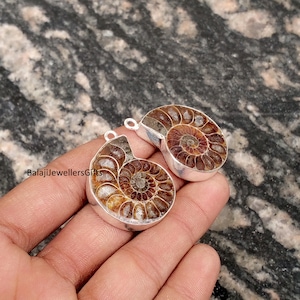 May include: Two fossilized ammonite pendants with silver settings. The pendants feature a spiral pattern in shades of brown and tan. The text "BalajiJewellersGifts" is visible on one pendant.
