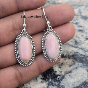 Peut inclure: Boucles d'oreilles pendantes en argent avec des pierres précieuses ovales rose pâle. Les pierres sont serties dans un cadre argenté perlé. Les boucles ont un design simple à crochet. Le texte "BalajiJewellersGifts" est visible.