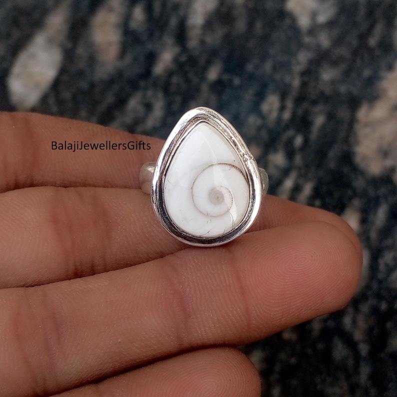 May include: A silver ring with a white, teardrop-shaped stone, displaying a spiral pattern. The stone is set in a silver bezel. The ring is shown on a hand. The text "BalajiJewellersGifts" is visible.