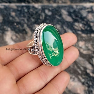 May include: A silver ring featuring a large, oval-shaped, glossy green gemstone. The ring has an ornate, filigree design around the gemstone and on the band. The text "BalajiJewellersGifts" is visible on the ring.
