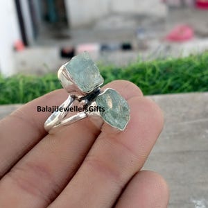 May include: A silver ring featuring two rough-cut, pale blue-green aquamarine stones. The ring has a simple band and the text "BalajiJewellersGifts" is visible on the ring.